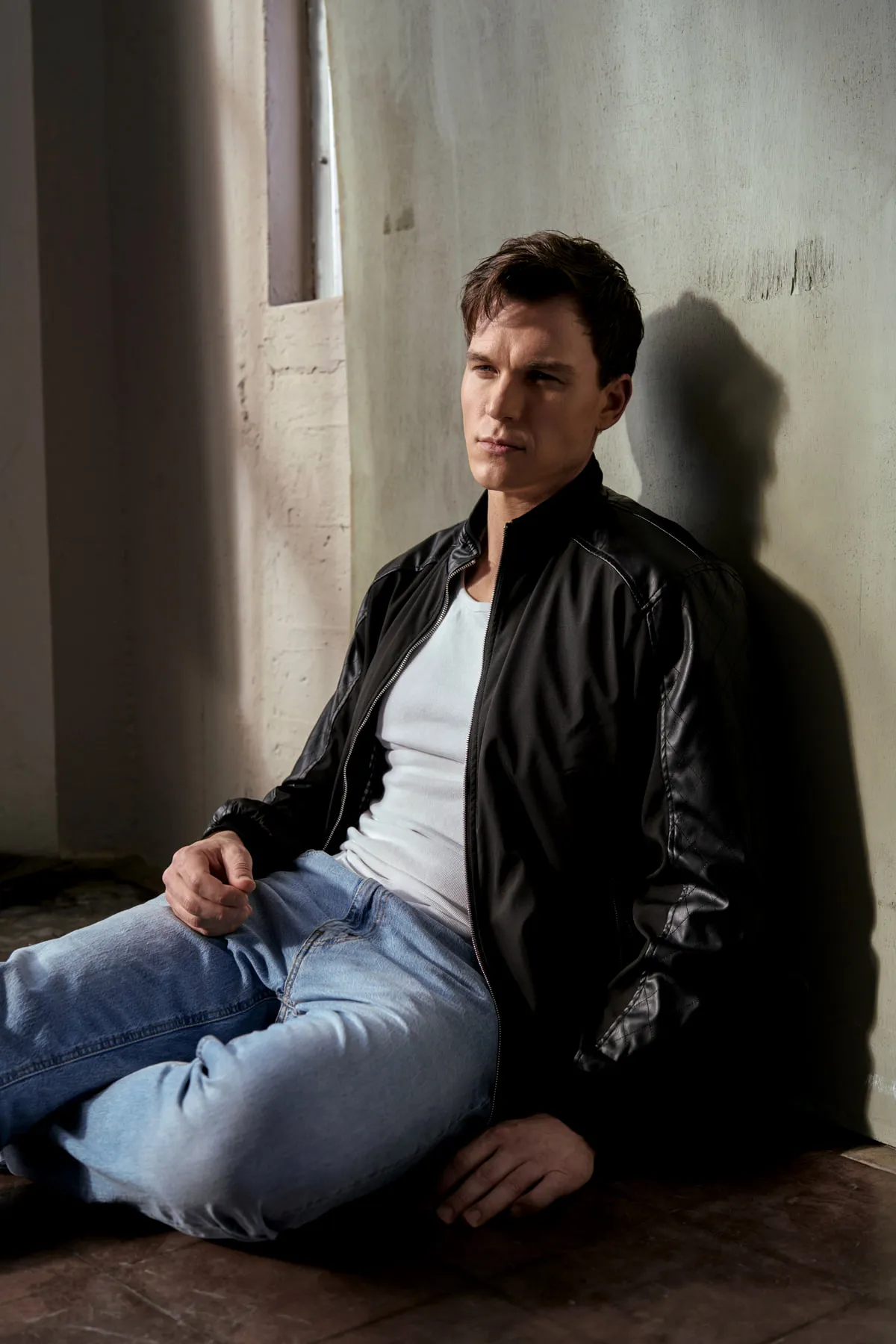 Steven Roderick Jurich seated against a sunlit wall in a leather jacket and jeans