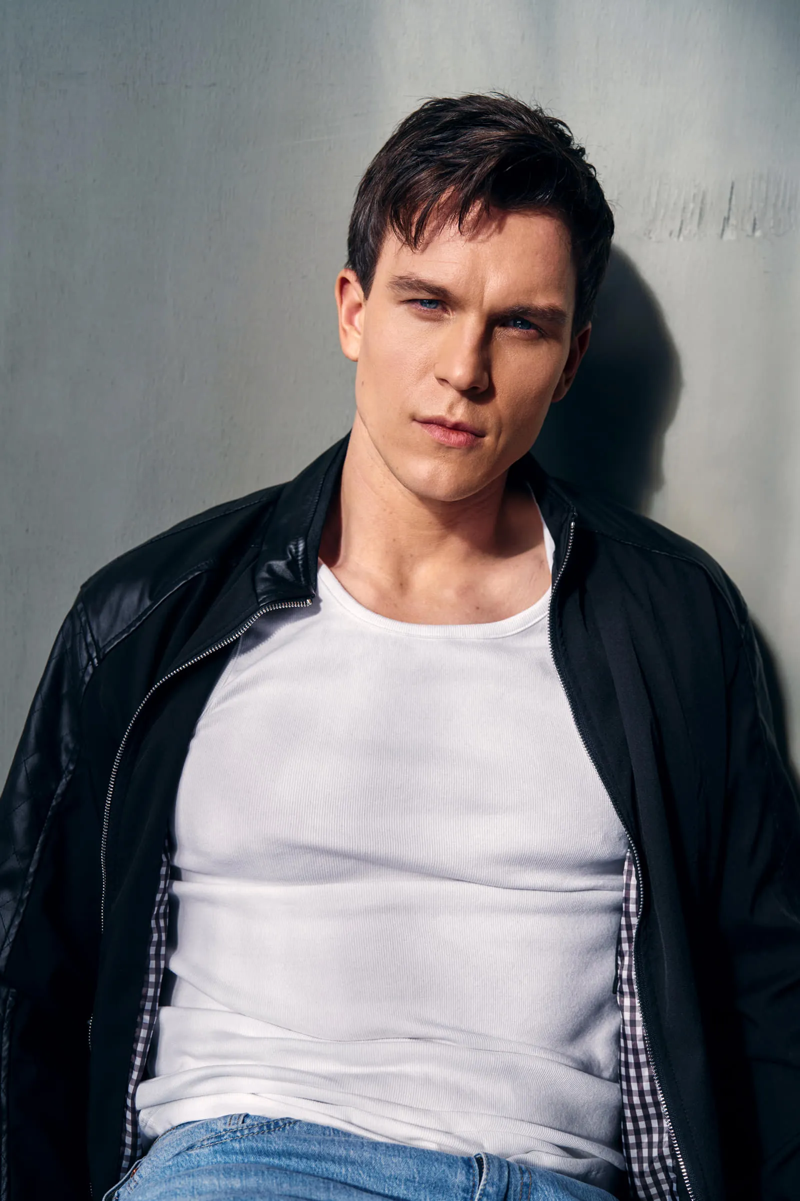 Steven Roderick Jurich against a sunlit wall in a leather jacket over a white tee